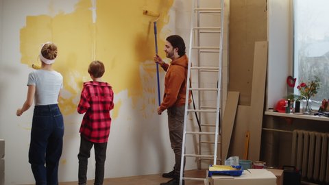 Family painting walls in yellow, making overhaul together with child. Husband and wife doing renovating with kid son. Interior design, new apartment. Boy helping to his parents. 