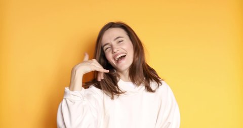 Friendly looking positive straight hair woman shows shaka sign gestures, tongue out indoor being in high spirit, wears white hoody isolated on yellow background. Hang loose!