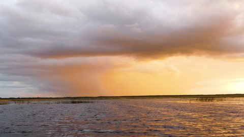 Rainy and windy cloud creeps on lake like snake, front of cloud system along coastline. Thunderstorm menacingly illuminated by setting sun, prestorm environment (thundery and oppressive air)