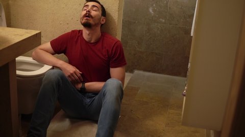 Young sick guy is sitting on the floor of bathroom next to toilet and holding his belly. Caucasian man feels nausea after food poisoning. He is suffering from stomach and head ache and wants to vomit.