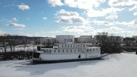 Steamboat docked for winter and closed up. Tarp over windows on main level of boat. River iced over with snow. Bright blue sky with fluffy white clouds. 