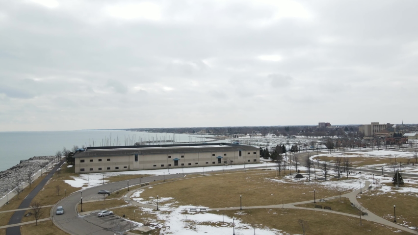 Aerial view of Lake Michigan. Park with sidewalk and road circling. Statue in park. Parking lot. Vehicles driving by. Cloudy gray sky. 