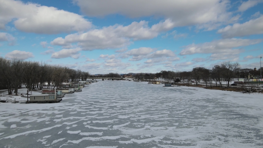 Winter view of river in Wisconsin. Swirling snow on the ice. Boat houses and trees line the shorelines. Boat houses boarded up for the season. 