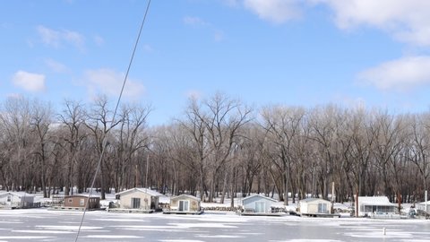 View of houseboats across river in Wisconsin in winter. Blue sky and fluffy clouds. Houseboats boarded up for season. Snow swirls on the ice. 