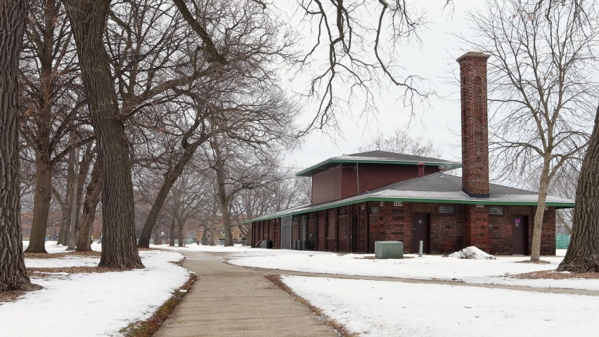 City park shelter in Kenosha, wisconsin, after light snowfall. Sidewalks cleared of snow. Bare branches. A few dry leaves scattered on the ground. Bike rack empty outside of the building. 