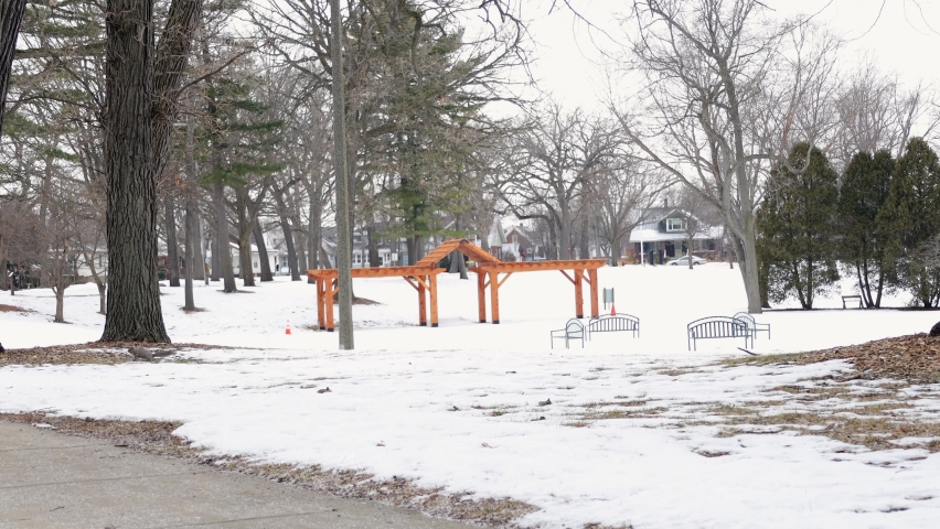 View of park in winter in Wisconsin. Bare trees in the park with a bit of grass peaking through the snow. Sidewalk for strolling. Playground equipment for playing. 
