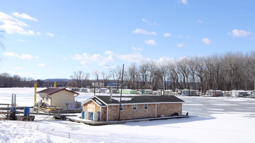 Large houseboat boarded up on the frozen river in Wisconsin. Houseboats lining the shoreline. Park with forest across the water. Blue sky with fluffy clouds. Wooden docks around the homes. 