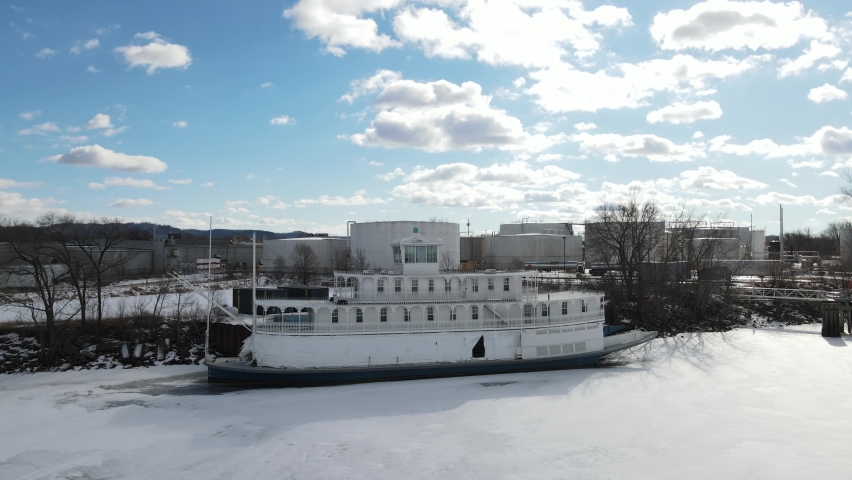 River boat closed up for the winter with tarps. Boat docked in the water with ice present on the water and snow. Industrial region seen in background.