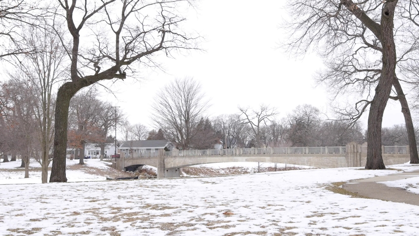 City park Kenosha wisconsin, walking bridge from park with sidewalk. Bare tree branches in the winter. Steep bank from creek. Gray cloudy sky. 