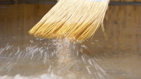 LOW ANGLE, CLOSE UP: Person uses a straw broom to sweep the flooded floor after an episode of heavy rain. Broom with rough straw bristles sweeps away the dirty water covering the basement floor.