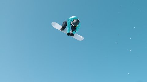 SLOW MOTION, BOTTOM UP: Athletic snowboarder flying through the air and doing a 360 grab on a sunny winter day in the Slovenian mountains. Spectacular shot of a snowboarding pro doing awesome tricks.