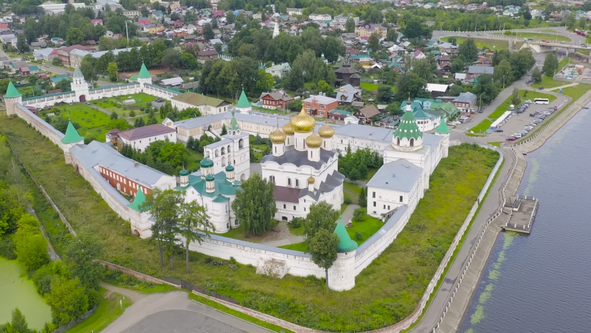 Russia, Kostroma. The Holy Trinity Ipatievsky Monastery, a historic Orthodox monastery with white stone walls, green conical towers, and a central cathedral crowned with five gilded onion domes, sits on a high riverbank at the confluence of the Kostroma and Volga rivers. The aerial hyperlapse captures the monastery