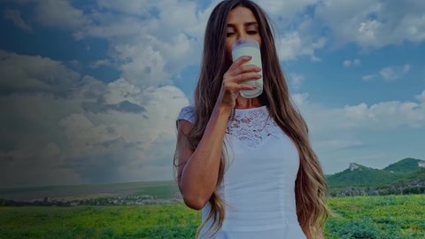 Beautiful woman in a white dress drinks milk from a glass against the backdrop of a green field and mountains.Portrait of a young girl drinking milk. Attractive lady enjoying a glass of milk the field