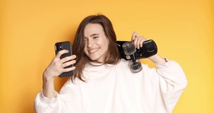 Stylish young happy girl in white hoody smiling while holding skateboard on shoulder and talk use mobile phone isolated on yellow background. Enjoying life. Healthy and careless lifestyle.