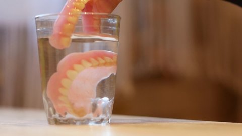 Close-up of a glass of water into which false teeth are immersed in dentures. An elderly person who has lost his teeth. Dental implantation clinic. Topic of tooth loss and prosthetics. selective focus