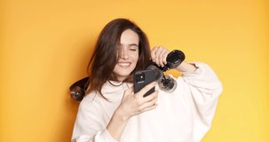 Stylish young happy girl in white hoody smiling while holding skateboard on shoulder and talk use mobile phone isolated on yellow background. Enjoying life. Healthy and careless lifestyle.