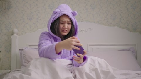 Upset millennial Asian woman playing video games on mobile phone and losing the game sitting on bed. Lady lost the game on smartphone. 