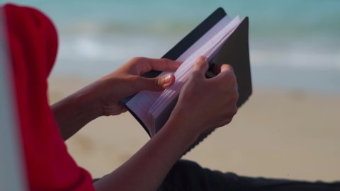 Close up shot of woman turning pages of book, in background of beach waves. Holiday concept
