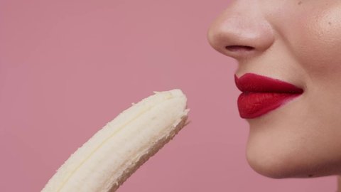 Close up. Sexy girl isolated on pink color background in studio eat banana. Pretty woman with red lips bite banana. Slow motion. Female eat banana