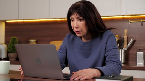 Exhausted aged woman uses laptop feeling worried reading bad news on social media. Stressed businesswoman works for computer frustrated thinking of money debt, sitting on chair at desk in home kitchen