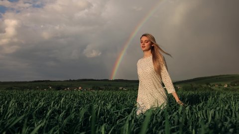 Smiling girl in a field with green wheat and rainbow. Young beautiful blonde woman in a white dress spinning in green field