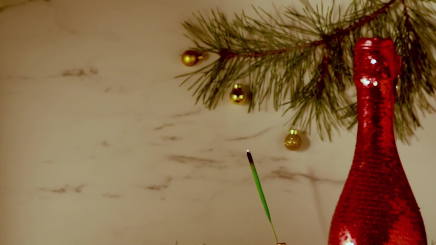 A burning sparkler on a light background and a bottle of champagne in red wrapping paper. Decorated with a spruce branch. Text space, copyspace
