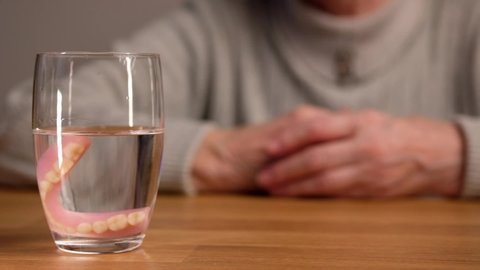 Elderly woman takes false teeth out of her mouth and puts it in glass of water. Oral, denture and dentures hygiene.