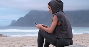 sporty female athlete looking his smartphone holding water bottle. an athletic thin girl in sportswear sits on a stone on the beach near the ocean with a phone, mountain, after a workout