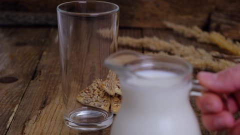 Female hand pouring vegetable milk from a jug into a glass on a wooden background with whole grain crackers or cookies