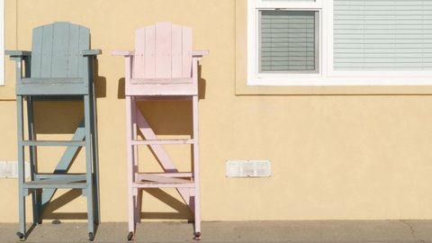 Two vintage wooden lifeguard chairs by yellow wall, California USA. Empty life guard retro high seat by ocean sea beach. Minimal simple trendy pop atmosphere, summertime aesthetic. Los Angeles vibes.