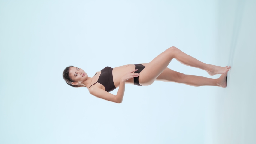 Vertical long shot of young slim good-looking Asian brunette woman with long ponytail in black underwear turns to the camera on tiptoes touching her hip on pale blue background | Leg skin care concept | Shutterstock HD Video #1084469959