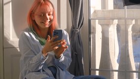 Happy young girl with dyed hair browsing social media on mobile phone. Emotional female with colored hair using modern smartphone and laughing out loud. 4K stock video of female with rainbow hairstyle