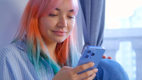 Young woman with dyed hair browsing social media news feed in cellphone. Cute girl with colored hair using mobile app on modern smartphone. 4K stock video of pretty white female with rainbow hairstyle
