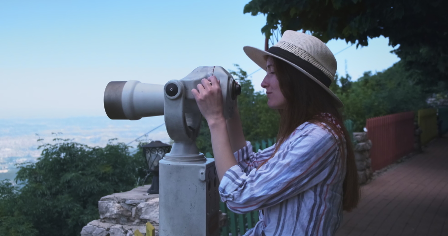 Happy woman traveler looking through viewing binoculars at Tirana city panorama. Caucasian woman in straw hat on viewpoint watching in telescope of sights observes summer cityscape view Daitit Albania