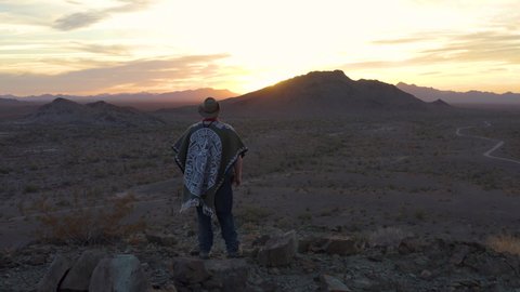 A poncho wearing wild west hero stands atop a southwestern mountain peak looking out upon a rugged desert landscape as the sun sets on the horizon. The camera makes an aerial dolly out motion.