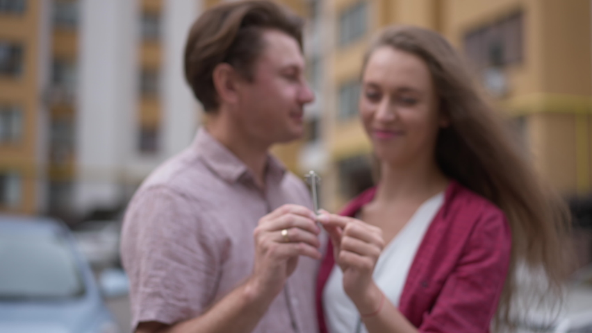 Close-up key in couple hands with blurred Caucasian man and woman talking smiling at background. Happy husband and wife buying new apartment posing outdoors in slow motion