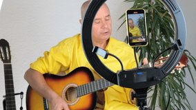 charming elderly man in a yellow shirt plays guitar in front of a ring light and a smartphone, teaches students online skills, records a training video, concept performance during covid 19