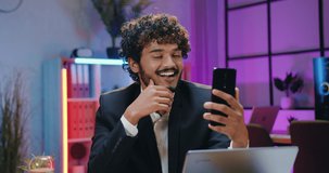 Handsome smiling successful bearded arabian businessman in suit enjoying phone video chat while overtime working in evening office,front view