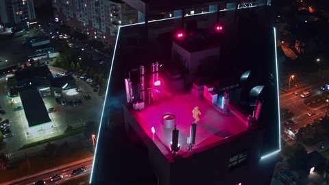 Aerial cityscape with artist performance. Young professional dancer performing contemporary breakdance on skyscraper rooftop. Neon party lights.