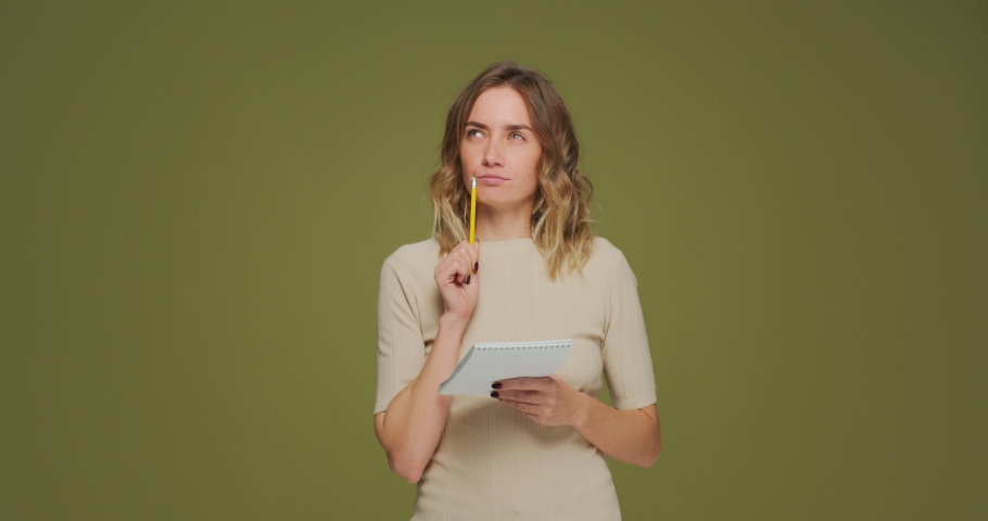 Pensive young woman thinking over smart idea, write plans, to-do list in notebook on green studio background
