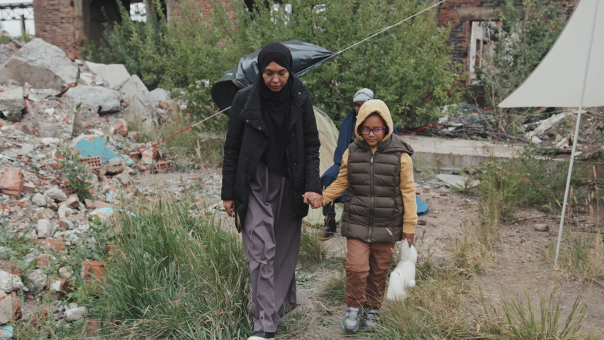 Tracking shot of biracial woman with daughter holding old stuffed toy walking along tents at refugee camp where other immigrants living on cold day