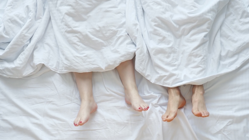 Young couple man and woman crosses legs stuck out of white sheet lying in soft bed in early morning in bedroom at home upper view.
