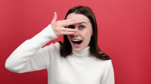 Portrait of young caucasian woman looks through fingers, peeks with curious face, having suspicious, wears white sweater, posing isolated over red color background in studio. Watches secrets concept