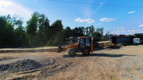 Use of heavy equipment in construction industry. Backhoe loader moves backwards on a construction site. Driver behind wheel of the excavator in blue work clothes. Site is covered with sand and gravel