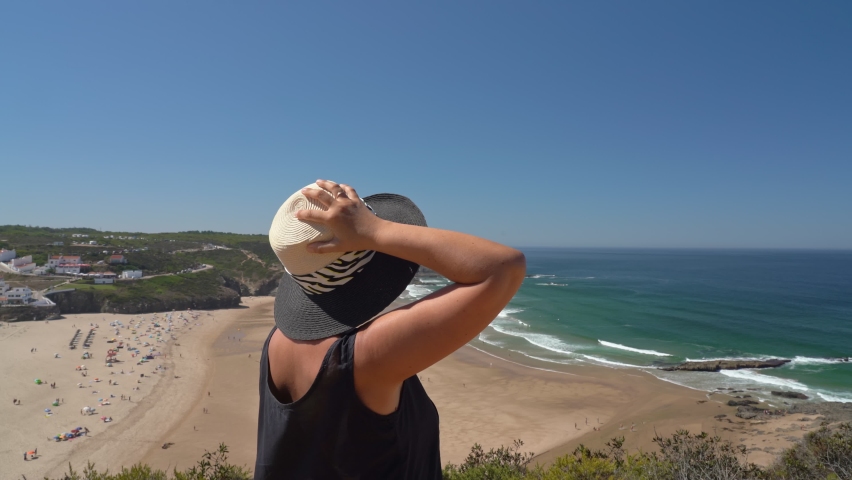 Woman in a middle-aged hat stands on a mountain slope and admires the beautiful view of the Portuguese beach Odeceixe in summer with tourists at the sea.