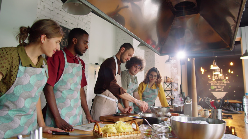 Zoom in of male chef adding alcohol to pan and flambeing food during cooking master class as group of multiethnic students watching flames with excitement Royalty-Free Stock Footage #1084535584