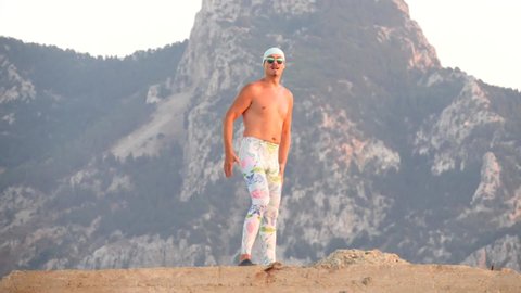 Young bald freak man tight leggings, topless, bandage on his head, with naked torso,stands at sunset, takes off his glasses and looks at the sun. Humor, unusual people. Slow motion video.