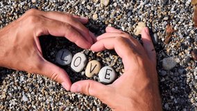 Men's hands folded in shape of a heart hold sea stones with word love written on stones. Letters are written on stones sea. Slow motion video. Text word love on stones. Valentine's Day, romance.
