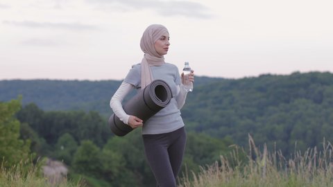 Side view of pretty young woman in hijab and activewear drinking water from bottle while standing at green park. Muslim female relaxing and refreshing after yoga practice outdoors.