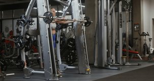 Young beautiful woman of athletic physique, dressed in a sports uniform, squats with a barbell on the exercise machine, doing in the gym. Video filming from the side, general plan of the gymnasium
